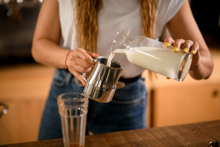 womans hand holds steel jug and pours milk into it from bottle.の写真素材
