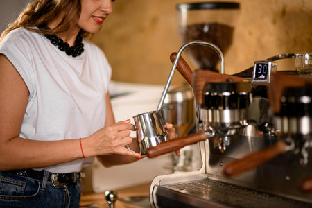 woman barista holds steel jug and makes whipped cream using coffee machine.