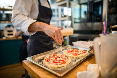 pizzas lie on baking sheet and male chef gently sprinkles grated cheese on itsの写真素材