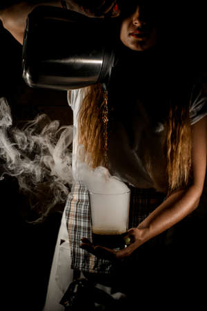 female barista pours drink from coffee pot into transparent steaming glass in her handの写真素材
