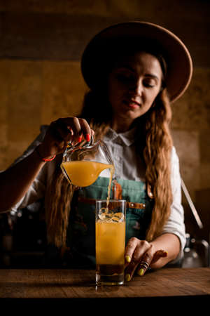 female bartender prepares drink and pours orange juice into glass with ice cubesの写真素材