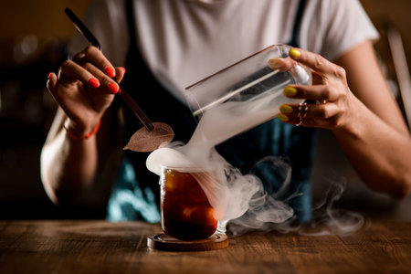 close-up of glass of drink from which bartender takes off flask and releases white smoke