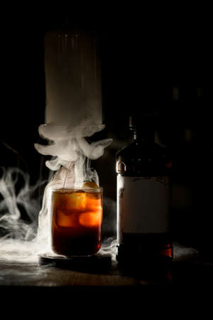 charming view of old-fashioned glass of cocktail and bottle in dark with white smoke aroundの写真素材