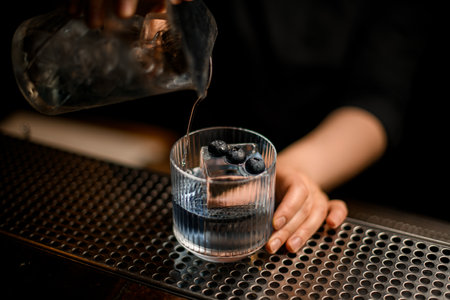 hand of woman bartender holds glass with ice and blueberries and pours into it cocktail from mixing cupの写真素材