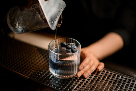 hand of female bartender holds old-fashioned glass with ice and blueberries and pours cocktail into itの写真素材