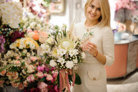 Smiling woman holds exclusive bouquet of different white flowers decorated with greenery in her handの写真素材