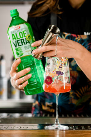 UKRAINE, KYIV - MARCH 12, 2021: Selective focus on beautiful glass with ice cubes in which hand pours liquid from steel jigger. Bottle of farmers aloe vera drink in hand of barmanのeditorial素材