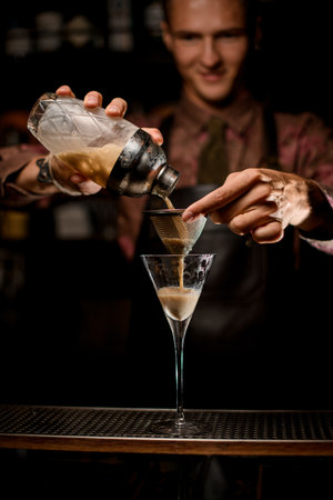martini glass and male hand holding sieve over it and gently pour liquid from shakerの写真素材