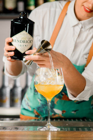 UKRAINE, KYIV - MARCH 12, 2021: beautiful glass with ice cubes stands on bar and bartender pours liquid from jigger into it and holds bottle of ginのeditorial素材