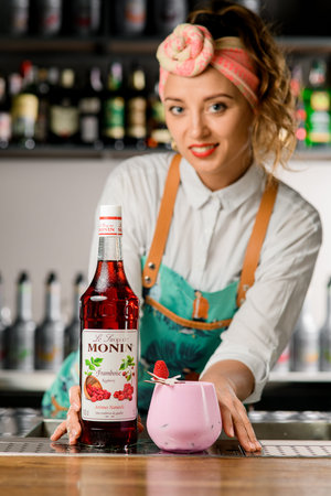 UKRAINE, KYIV - MARCH 12, 2021: smiling woman bartender holds bottle of raspberry syrup and glass of cocktail decorated with coconut and raspberryのeditorial素材