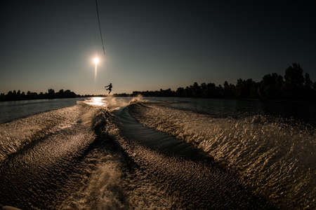 sportive man is jumping with wakeboard at trails behind boat. Wakeboarding on river. Summertime leisureの写真素材