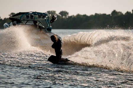 Athletic guy holds rope and balancing on wakeboard behind motor boat on splashing river waves.の写真素材
