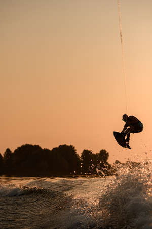 great view of active male rider holds rope and making jump on wakeboard at evening sky on background.の写真素材