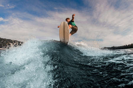 Active male wakesurfer jumping on a wake board on splashing river wave against skyの写真素材