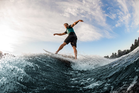 young man energetically balancing on wave on wakesurf board on splashing wave.