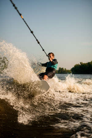 Athletic guy holds rope and speedly riding wakesurf board on splashing wave.の写真素材