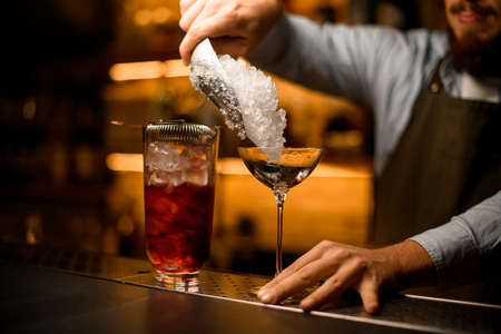 hand of barman holds empty glass on the bar and pours ice into it from a metal scoopの写真素材
