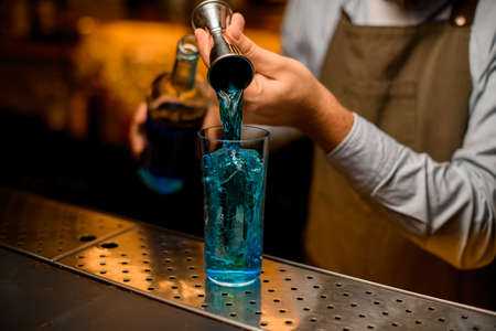 view of glass with ice in which the hand of bartender gently pours blue drink from a jiggerの写真素材
