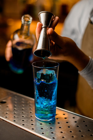 close-up of glass with ice in which the hand of bartender pours blue drink from a jigger