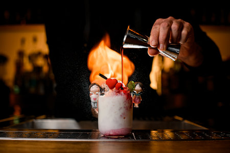 Bartender holds steel jigger in his hand and neatly pours syrup into glass with cocktailの写真素材