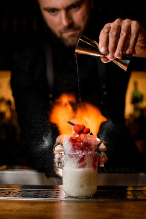 Bartender holds steel jigger in his hand and adding syrup into glass with cocktailの写真素材
