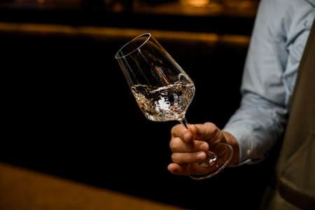 hand of man gently holds tilted transparent wine glass with a drinkの写真素材
