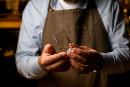 selective focus of tumbler glass in hands of male bartender in brown apron.の写真素材