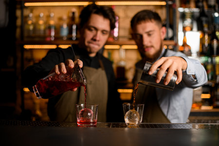 two glasses with ice in which bartenders carefully pour different cocktailsの写真素材