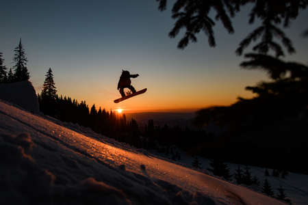 view of skier silhouette making jump in the air while sliding down snow-covered slope against the backdrop of sunsetの写真素材