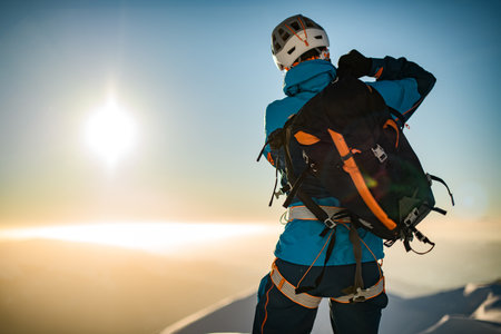 beautiful rear view of man in helmet and ski suit with backpack on his back against background of sky and sun. Copy space. Ski tour tourism conceptの写真素材
