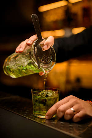 close-up of hand accurate holding mixing cup with cocktail and strainer and pours beverage into glassの写真素材