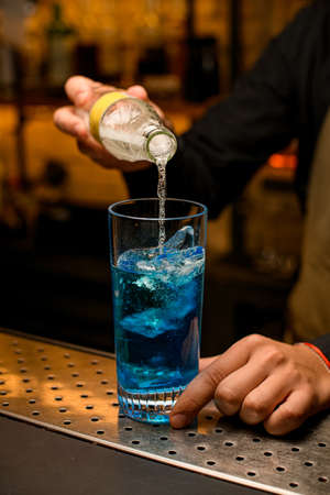 Close-up of glass with ice cubes and blue beverage into which bartender accurate pours drink from bottleの写真素材