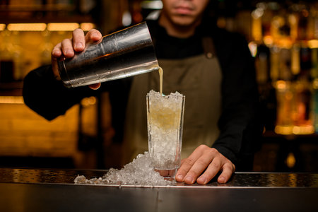 front view of glass full of ice pieces in which the hand of bartender pours a cocktail from shakerの写真素材