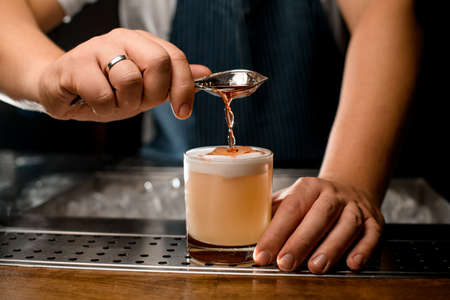 close-up of glass with drink and hand of barman pours liquid from spoon into itの写真素材