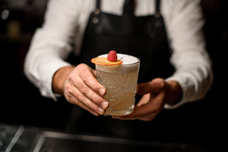 Close-up of glass with foamy cocktail decorated with orange slice and a raspberry in male handsの写真素材