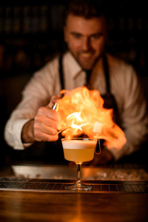 Bartender sprinkles on glass with cocktail on bar counter and makes fire flame over it.の写真素材