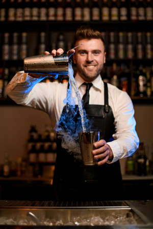 smiling male bartender skillfully pours a steaming cocktail from one shaker cup to anotherの写真素材