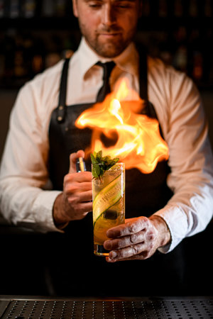 hand of man holds cocktail glass with cucumber slices and mint, sprinkles and carefully sets it on fireの写真素材