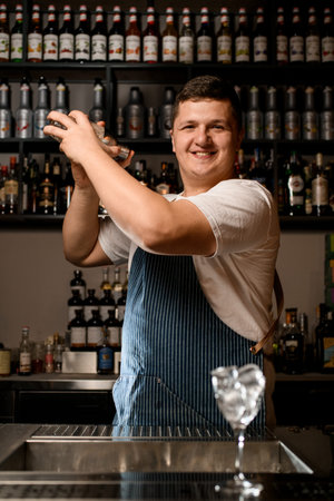 Bartender professionally holds by two hands glassy shaker with cold alcoholic cocktail and shakes...