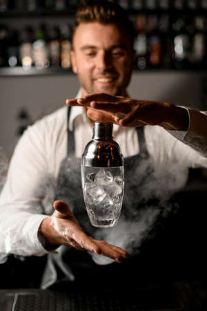 Young handsome barman in bar interior skillfully throws a shaker with ice into the airの写真素材