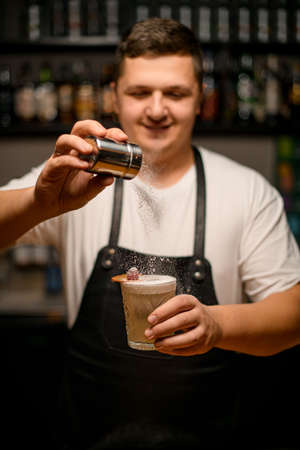 barman gently holds glass full of foamy cocktail and sprinkles on it with sugar powderの写真素材