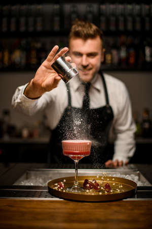 Selective focus on glass with pink foamy cocktail which hand of male bartender sprinkles it with sugar powder.の写真素材