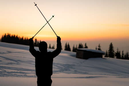 rear view of man with ski poles crossed over his head against the backdrop of sunset skyの写真素材