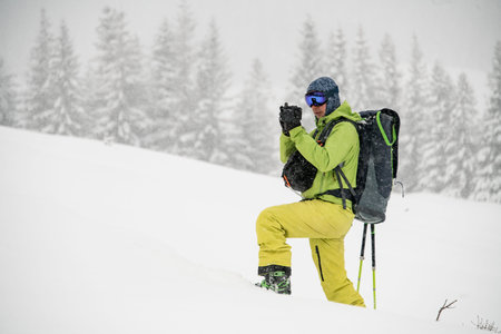 Side view of male skier in green and yellow ski suit with backpack standing alone and taking photo...