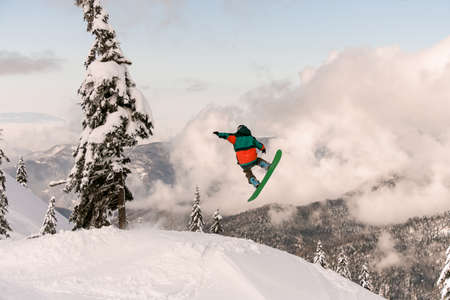 Snowboarder freerider jumping from snow-capped mountain slope on background of trees and cloudy skyの写真素材