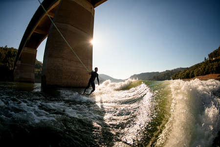 great view on guy in wetsuit holding rope with handle and riding on wakesurf board on wave.の写真素材