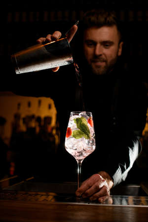 glass with ice cubes and bartender accurate pouring cocktail from shaker into itの写真素材