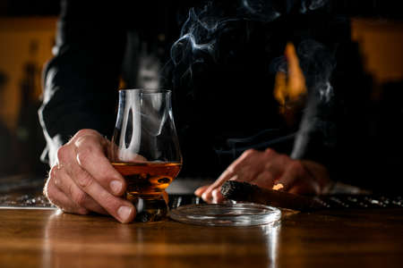 male hand holds glass with an alcoholic drink and ashtray with a smoking cuban cigar nearbyの写真素材
