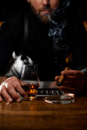 view on male hand holding glass with cognac and smoking cuban cigar over ashtrayの写真素材