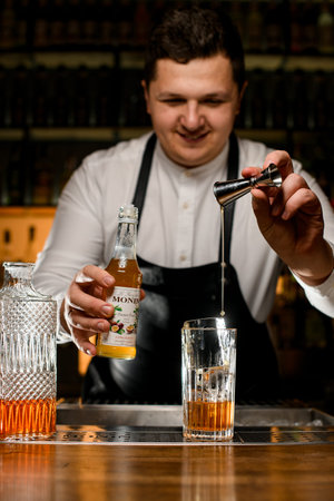 UKRAINE, KYIV - DECEMBER 06, 2021: bartender holds a bottle of monin syrup and pours liquid from a jigger into a glass with iceのeditorial素材
